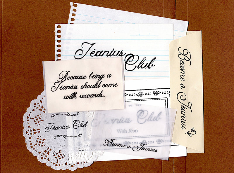 A flat-lay arrangement of vintage-style paper ephemera and stationery items on a brown textured background, including a lined notepad, cream cards with black script text reading 'Jeanius Club' and 'Become a Jeanius', a white lace doily, and a clear plastic sleeve.