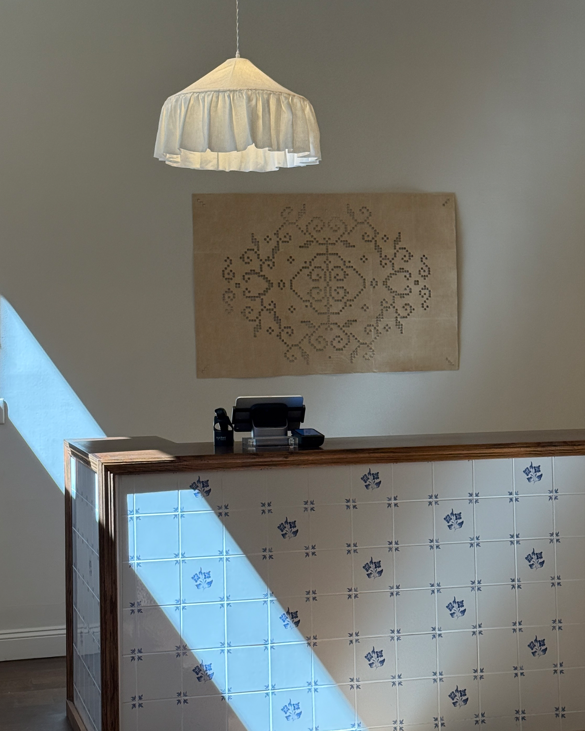 A modern interior scene featuring a wooden reception desk with blue and white patterned tiles, a beige wall hanging, and a white pleated pendant light casting a strong diagonal shadow on the wall.