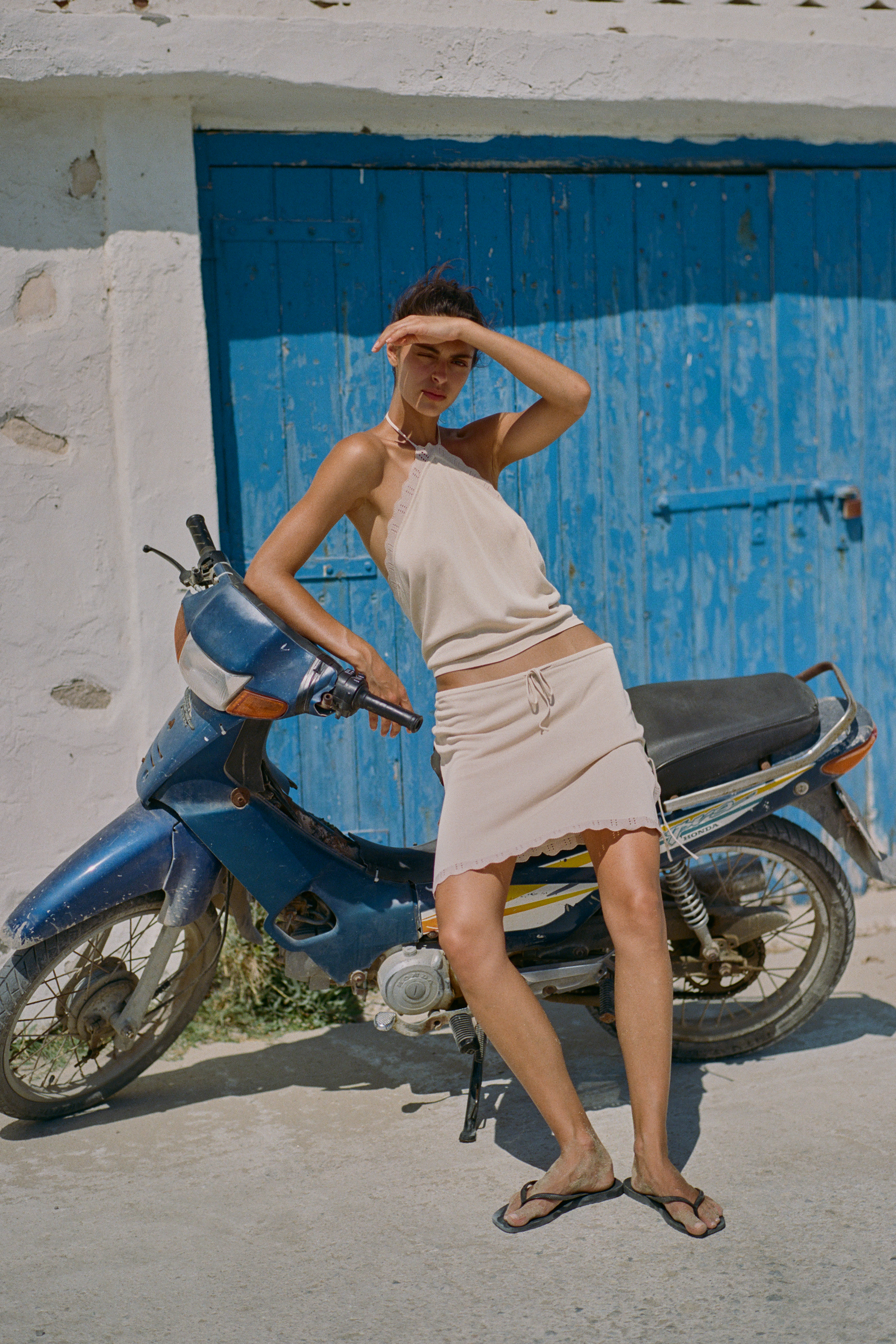 A woman posing outdoors next to a blue scooter in front of a weathered blue wooden door and white wall, wearing a light beige halter top and matching mini skirt
