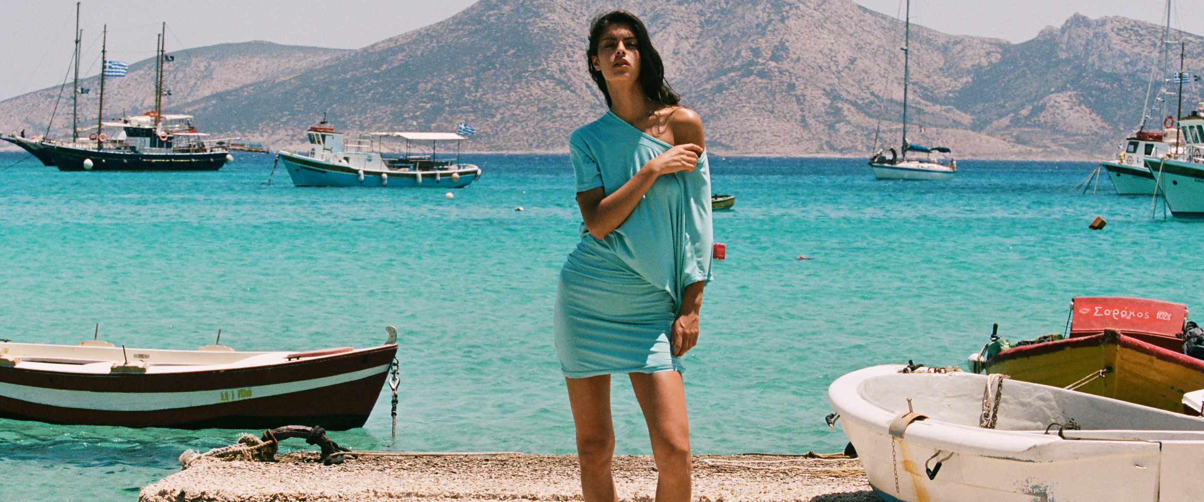 A woman wearing a light blue t-shirt dress stands on a rocky shoreline with turquoise water, boats, and mountains in the background.