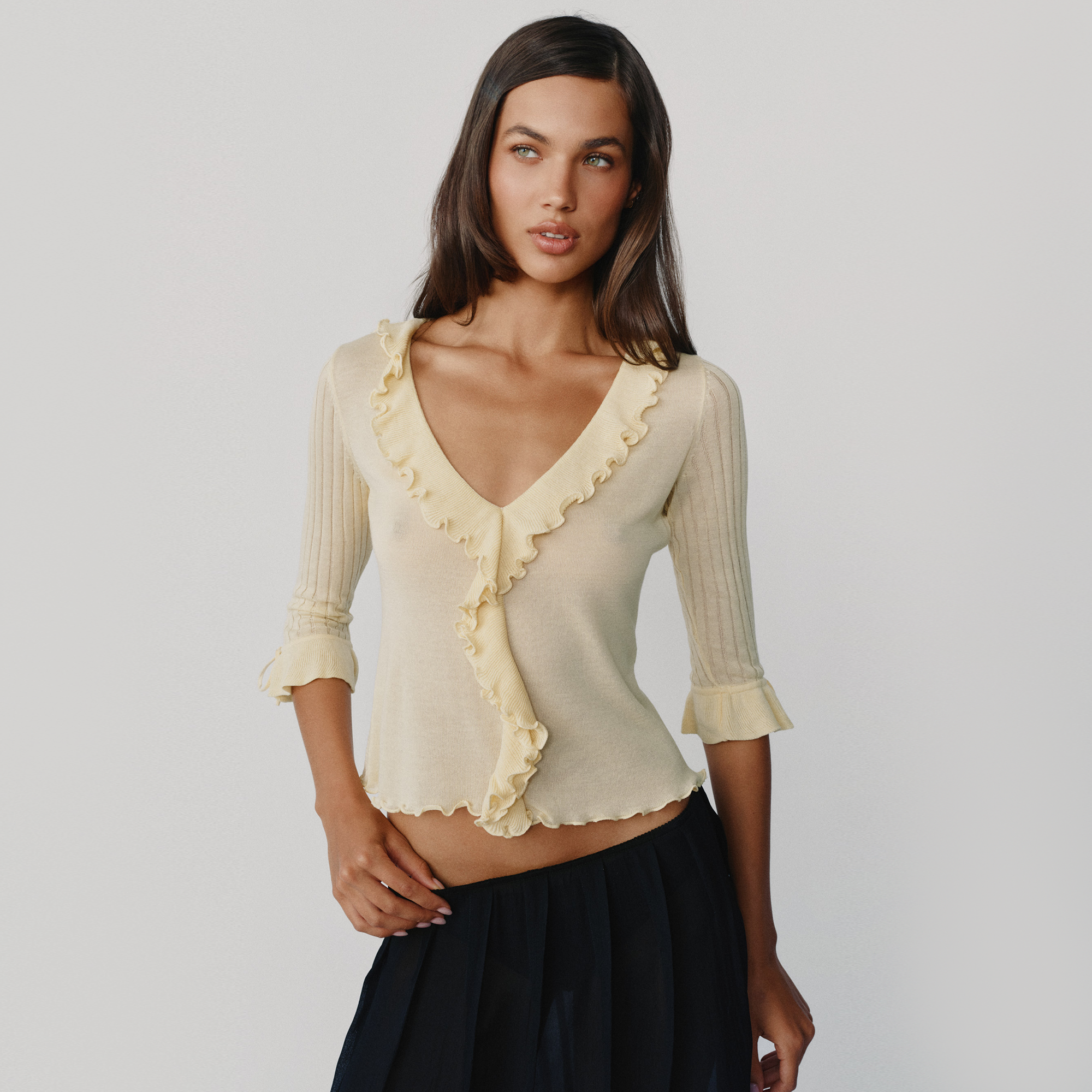 A woman modeling a pale yellow ribbed knit top with ruffle trim and three-quarter sleeves, paired with a dark skirt, against a light gray studio background.