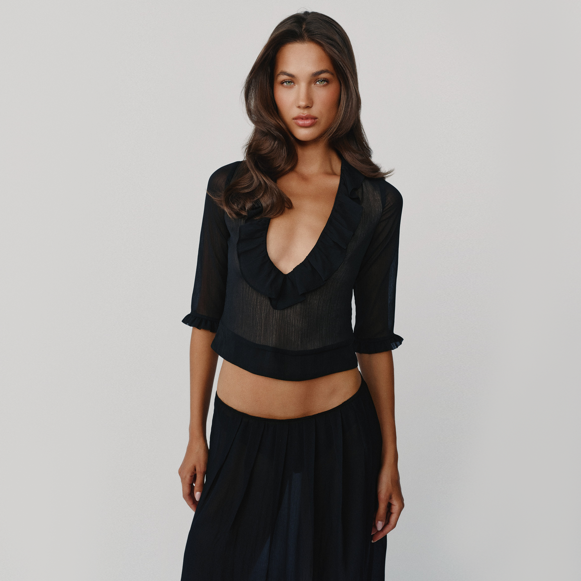 A woman modeling a black sheer mesh ruffle crop top with elbow-length sleeves and a black skirt against a light gray studio background.