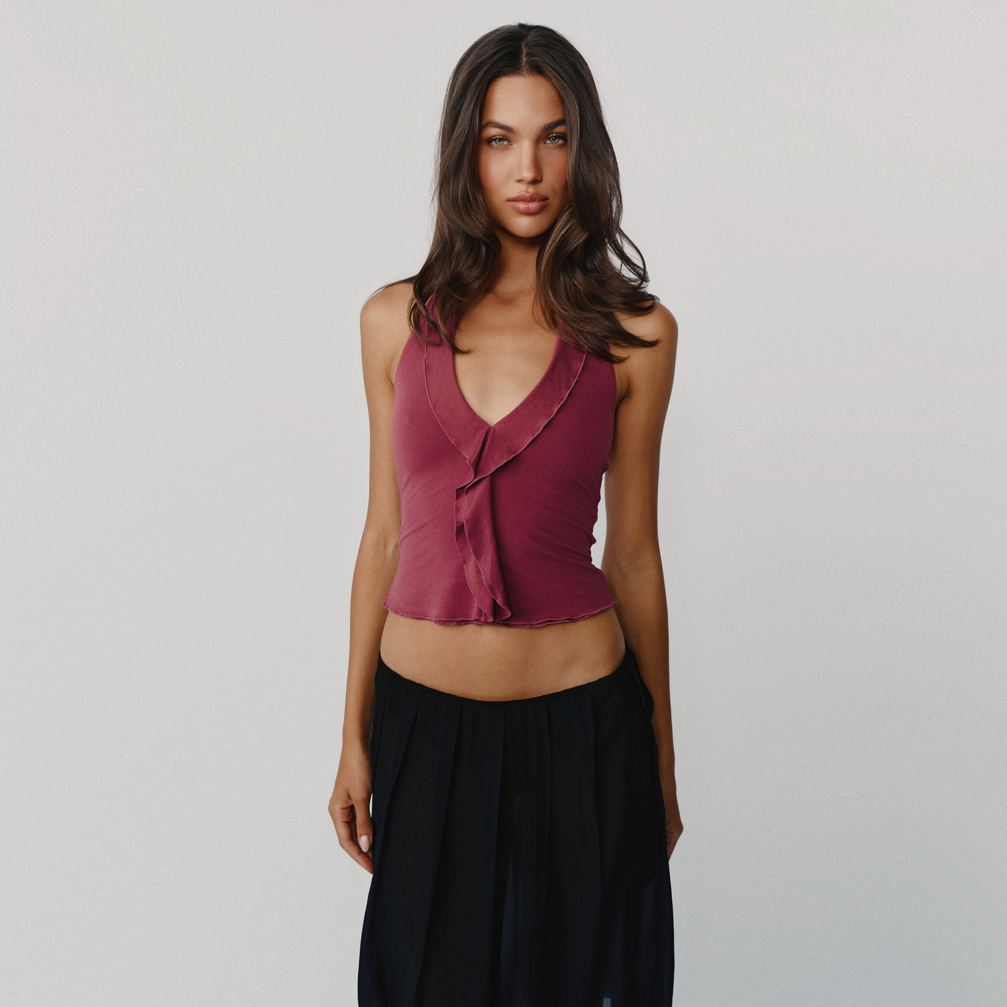 A woman modeling a sleeveless crop top in dusty burgundy, paired with black skirt against a plain light gray studio background.