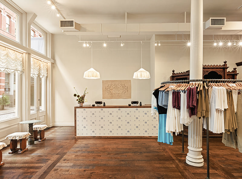 A boutique interior with a wood floor, white walls, hanging pendant lights, a patterned reception desk, and clothing racks filled with garments. No person is visible.
